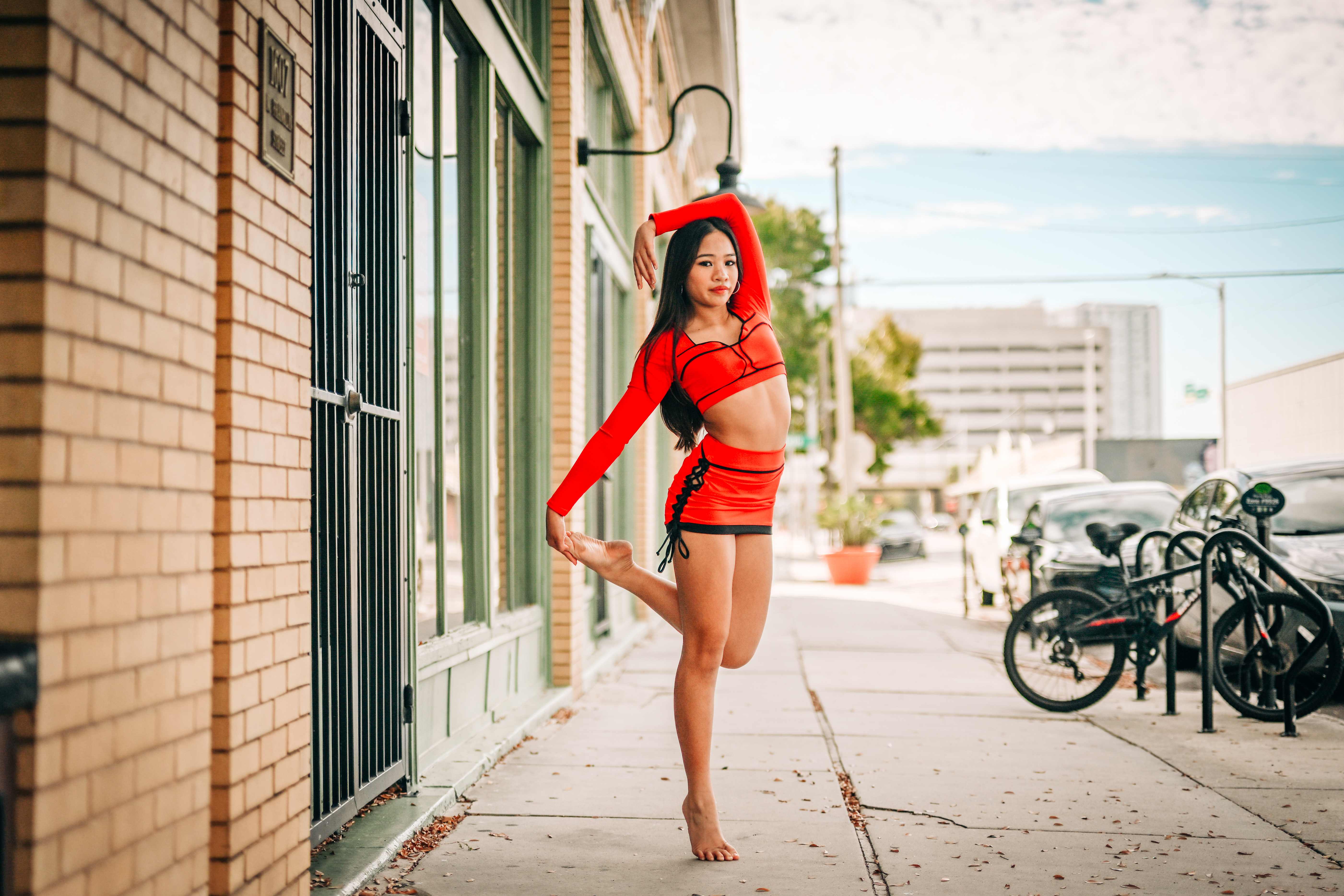 Richani balancing on one leg in a red outfit on a downtown sidewalk