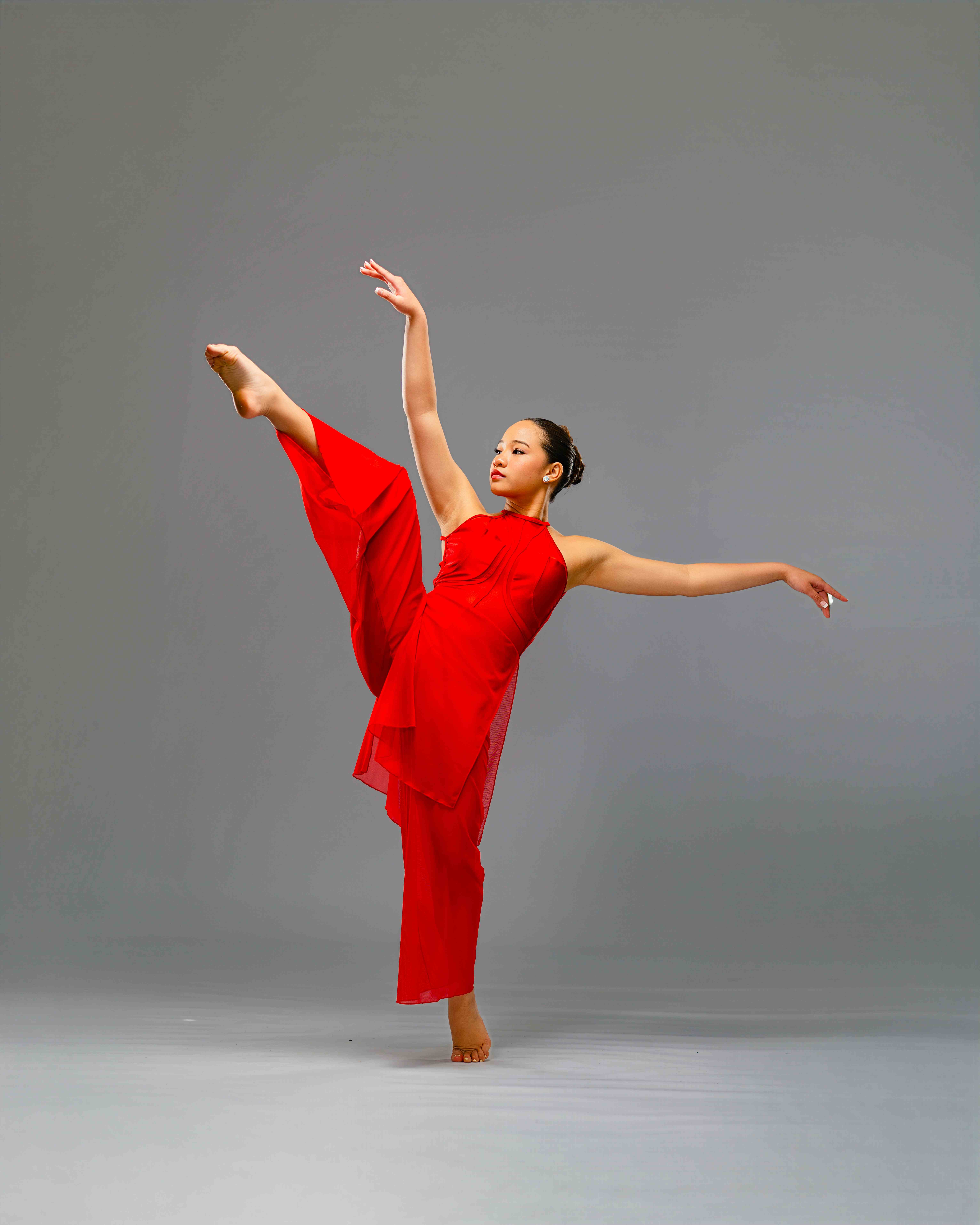 Richani Luong in a flowing red costume holding a high leg extension with arms outstretched in a grey studio
