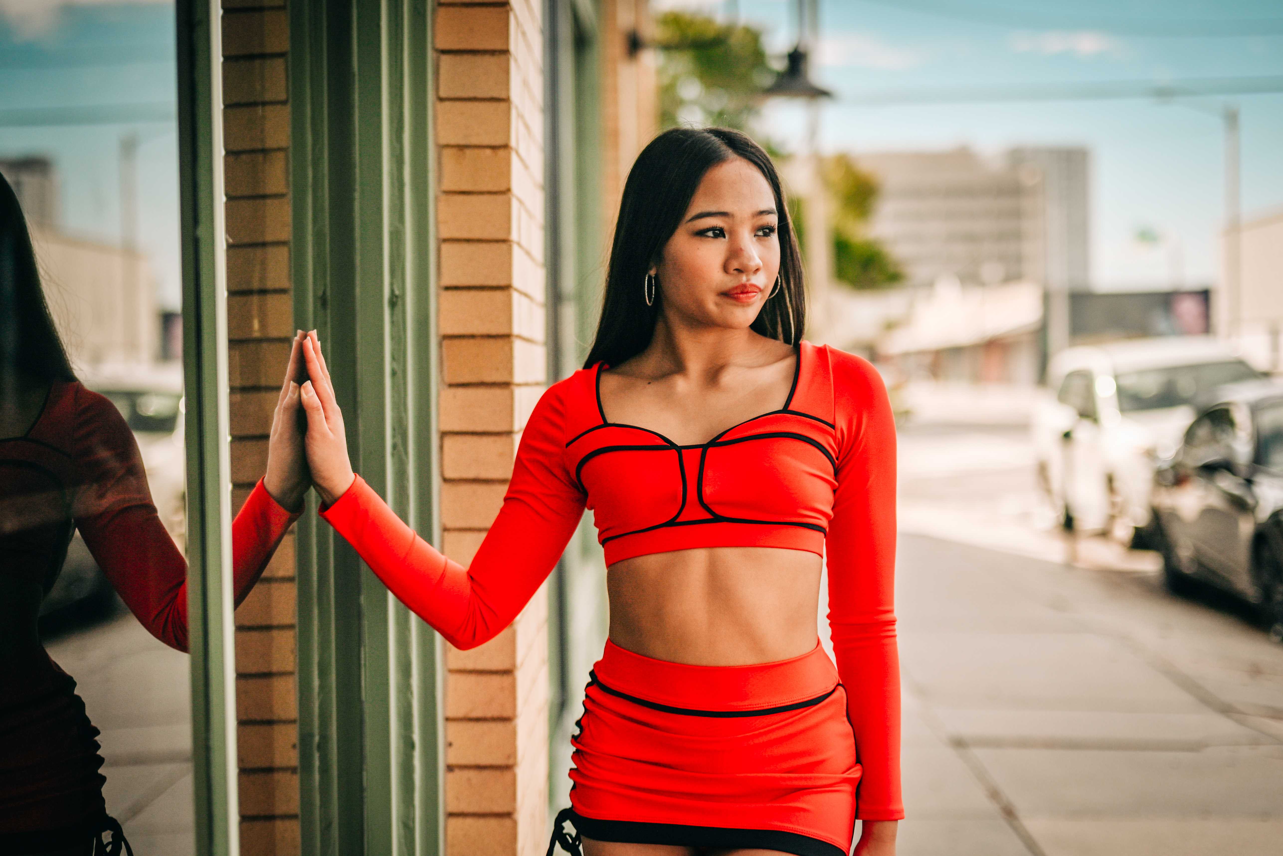 Richani in a red crop top touching her reflection in a storefront window