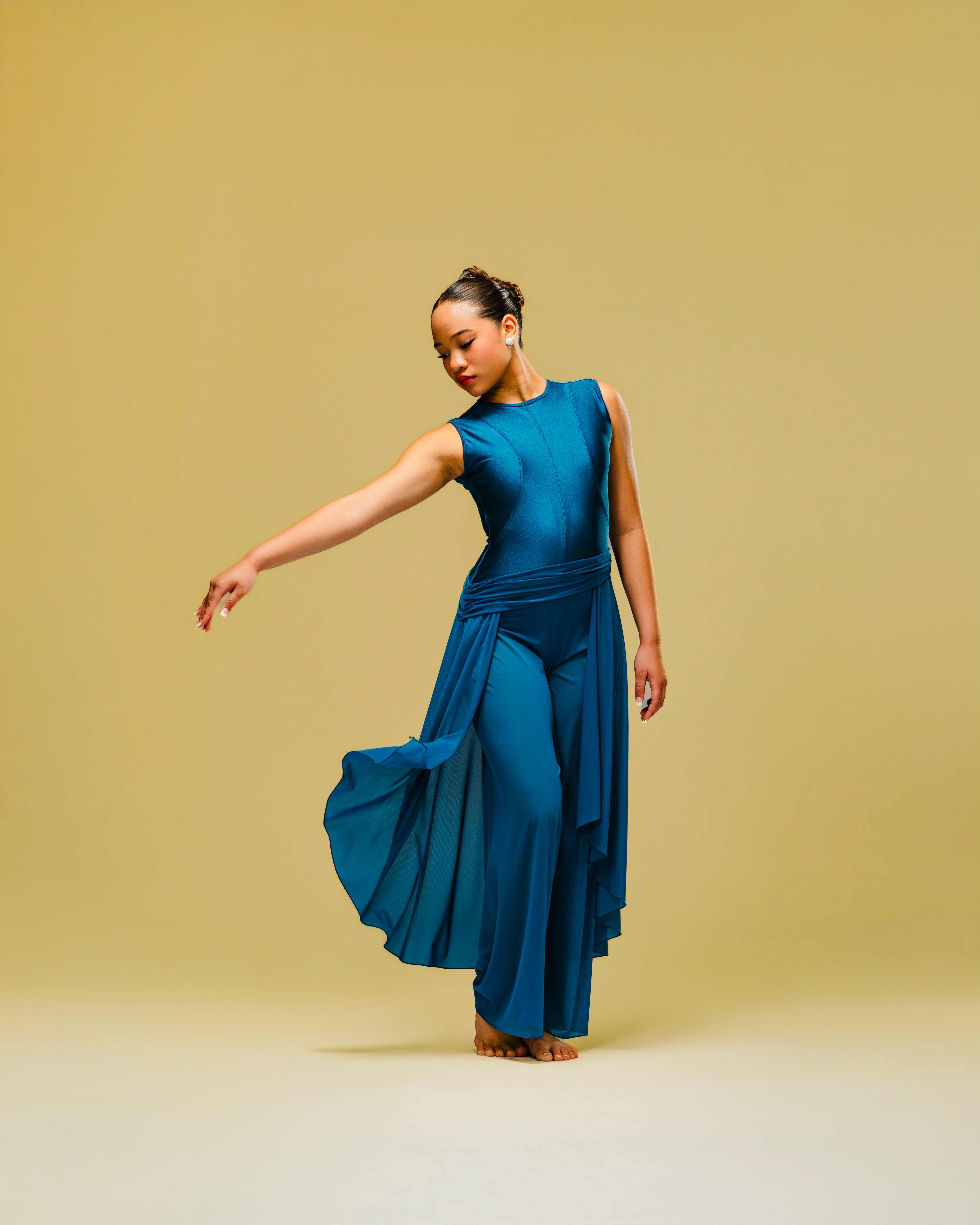 Richani in a teal jumpsuit with a flowing mesh overskirt, one arm reaching low as she looks down